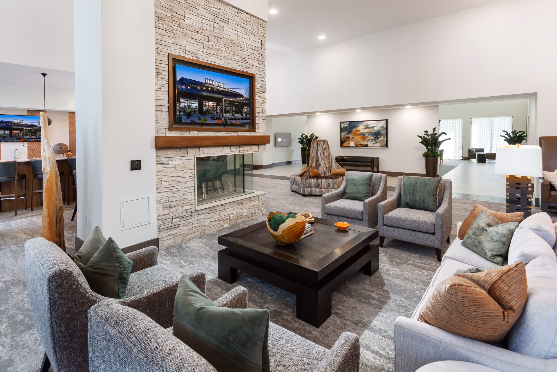 Bright modern communal living room with sofas and armchairs arranged around a central coffee table, a stone fireplace with a mounted TV, and decorative plants.