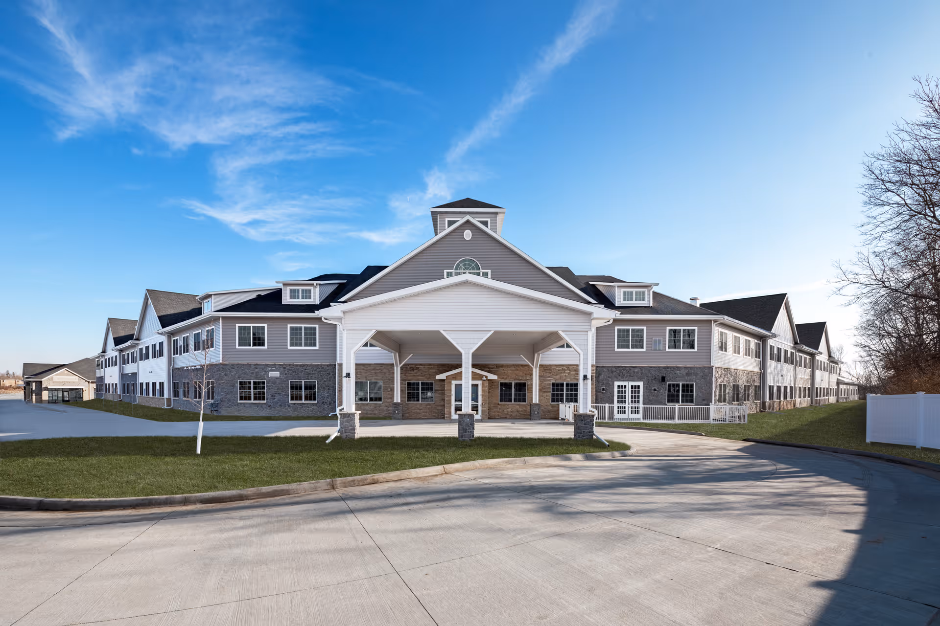 Front exterior of a large two-story assisted living building with a covered entrance, stone accents, driveway and lawn under a blue sky.