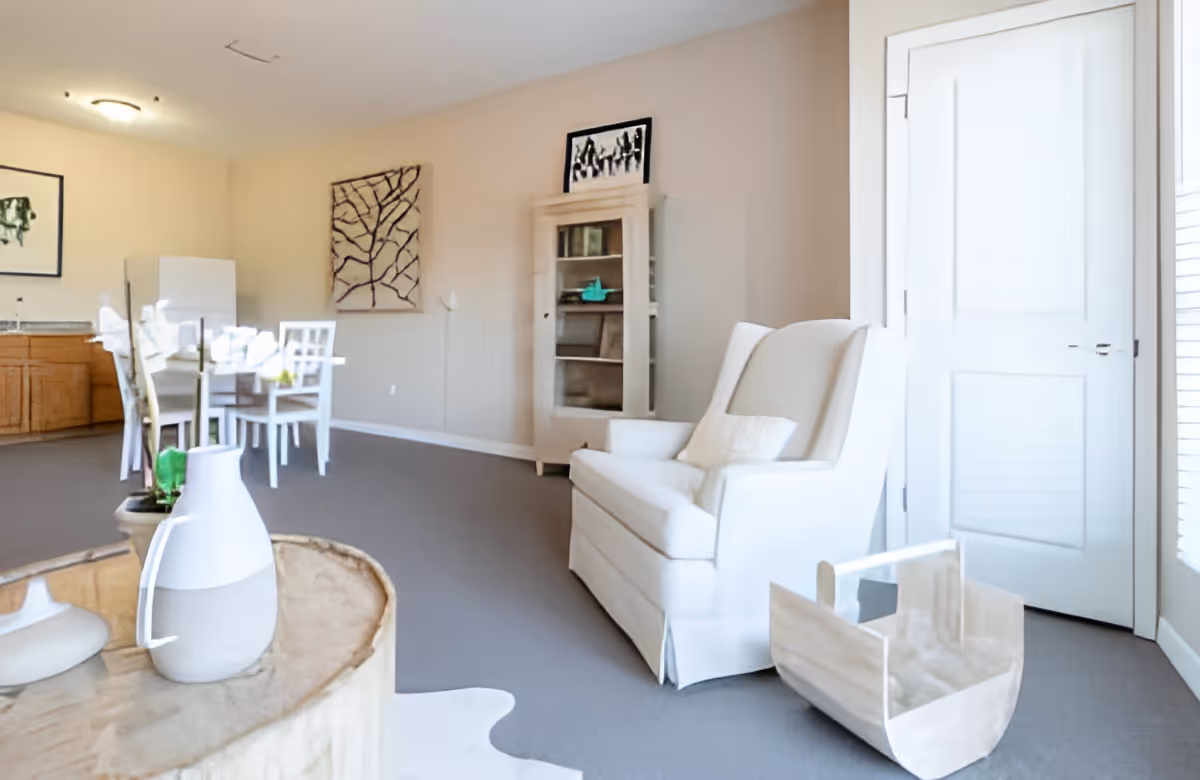 A bright and clean living area featuring a white armchair with a pillow, a wooden basket on the floor, a glass-topped round table with a white vase, and a dining area with a white table and chairs near a kitchen with wooden cabinets and a white refrigerator. The walls are beige with framed artwork, and there is a white door with blinds on the right side.