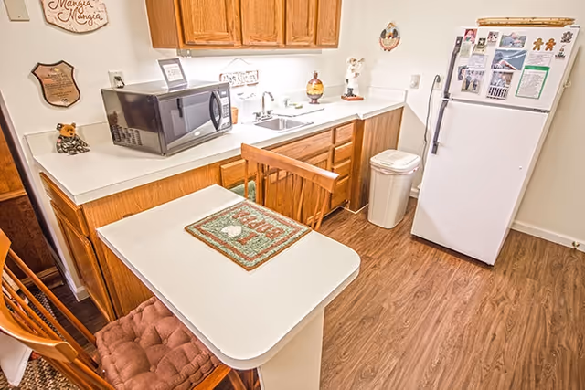 Small kitchenette with a white countertop and table, wooden cabinets, microwave, sink, refrigerator, and wooden chairs on a wood floor.
