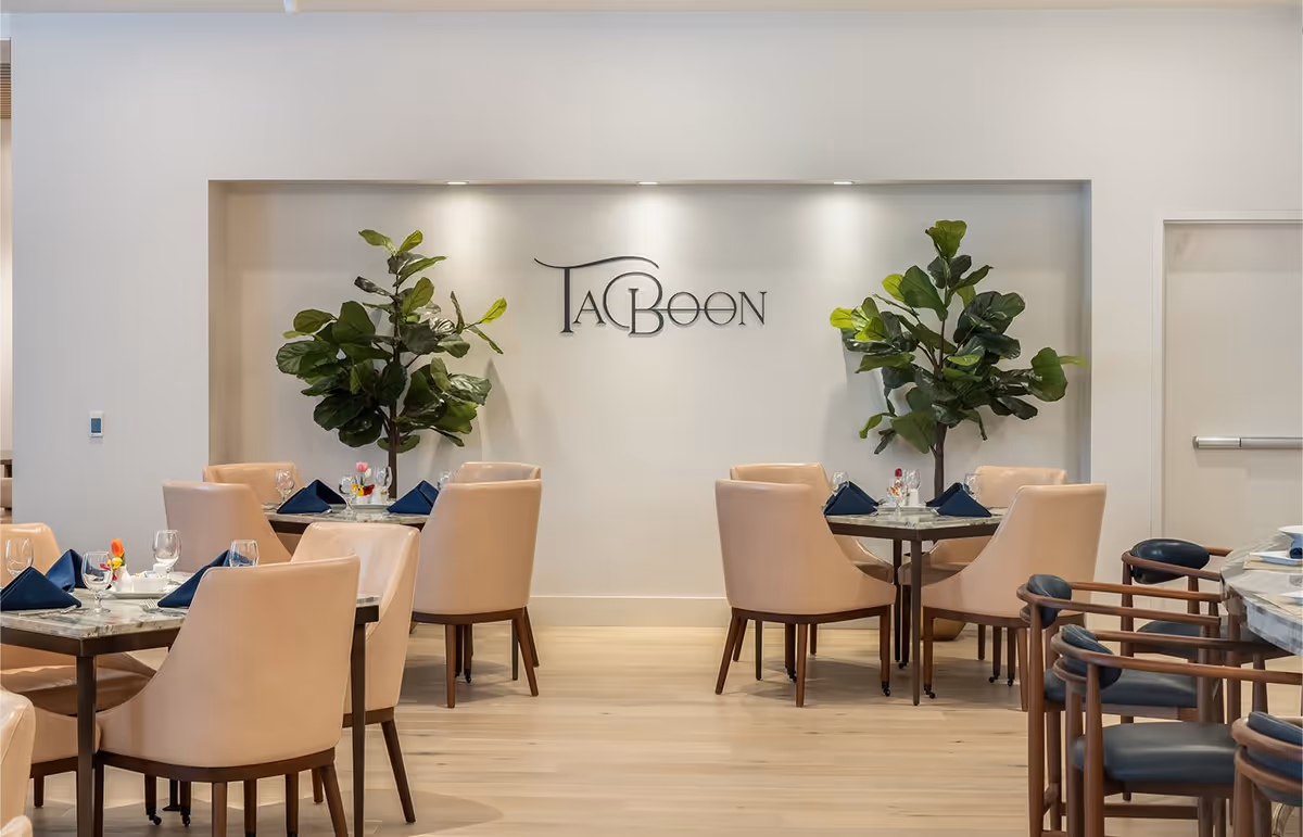 A modern dining room with beige upholstered chairs around tables set with glassware, plates, and navy blue napkins. Two large green plants flank a wall with the word 'TABOON' displayed in decorative letters. The room has light wood flooring and a clean, minimalist design.