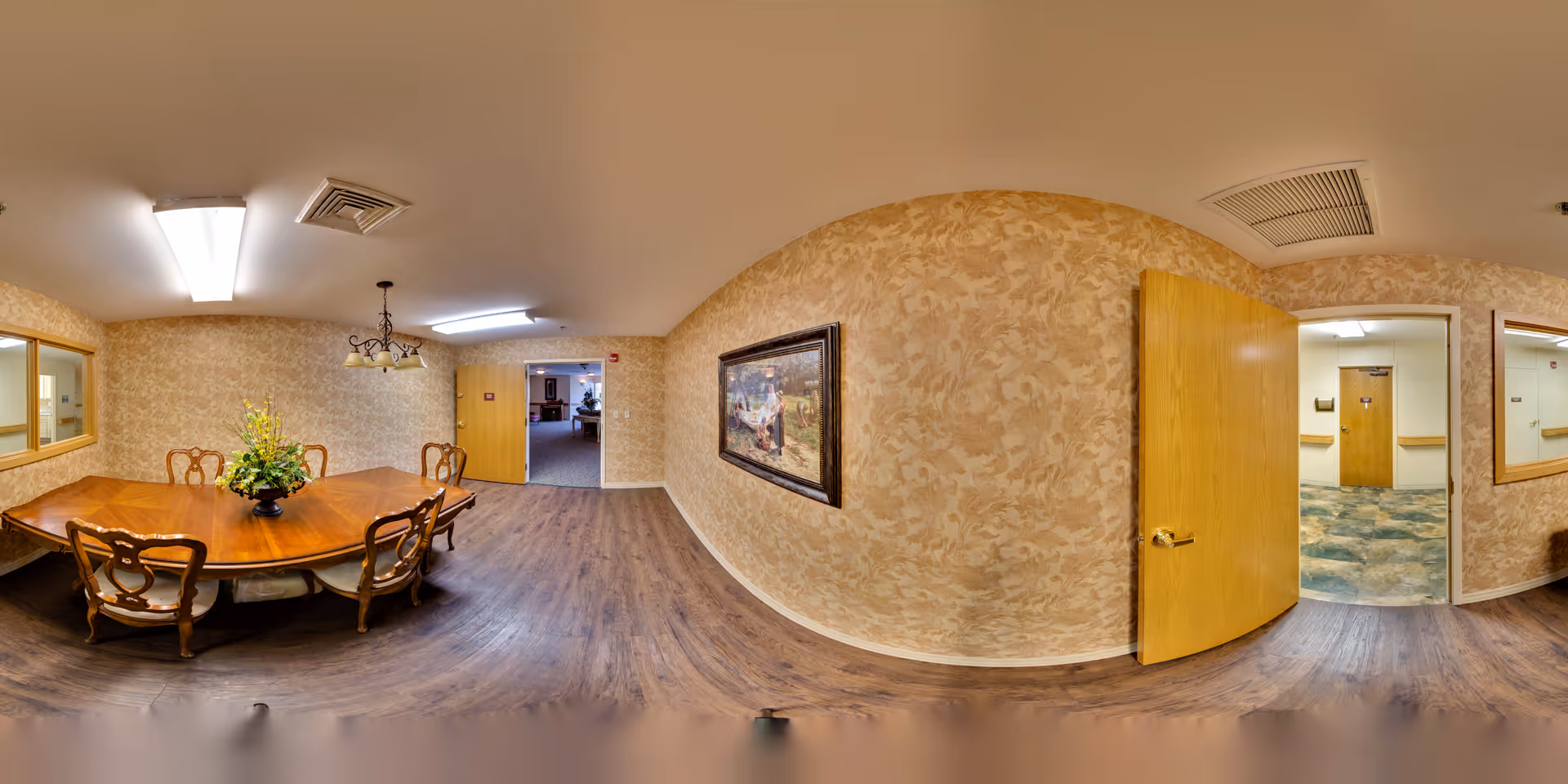 A dining room in Plantation Village Nursing Center featuring a wooden dining table with six chairs and a floral centerpiece. The room has patterned beige wallpaper, wood flooring, a chandelier above the table, and fluorescent ceiling lights. There are two open wooden doors leading to adjacent hallways, and a framed painting on one wall.