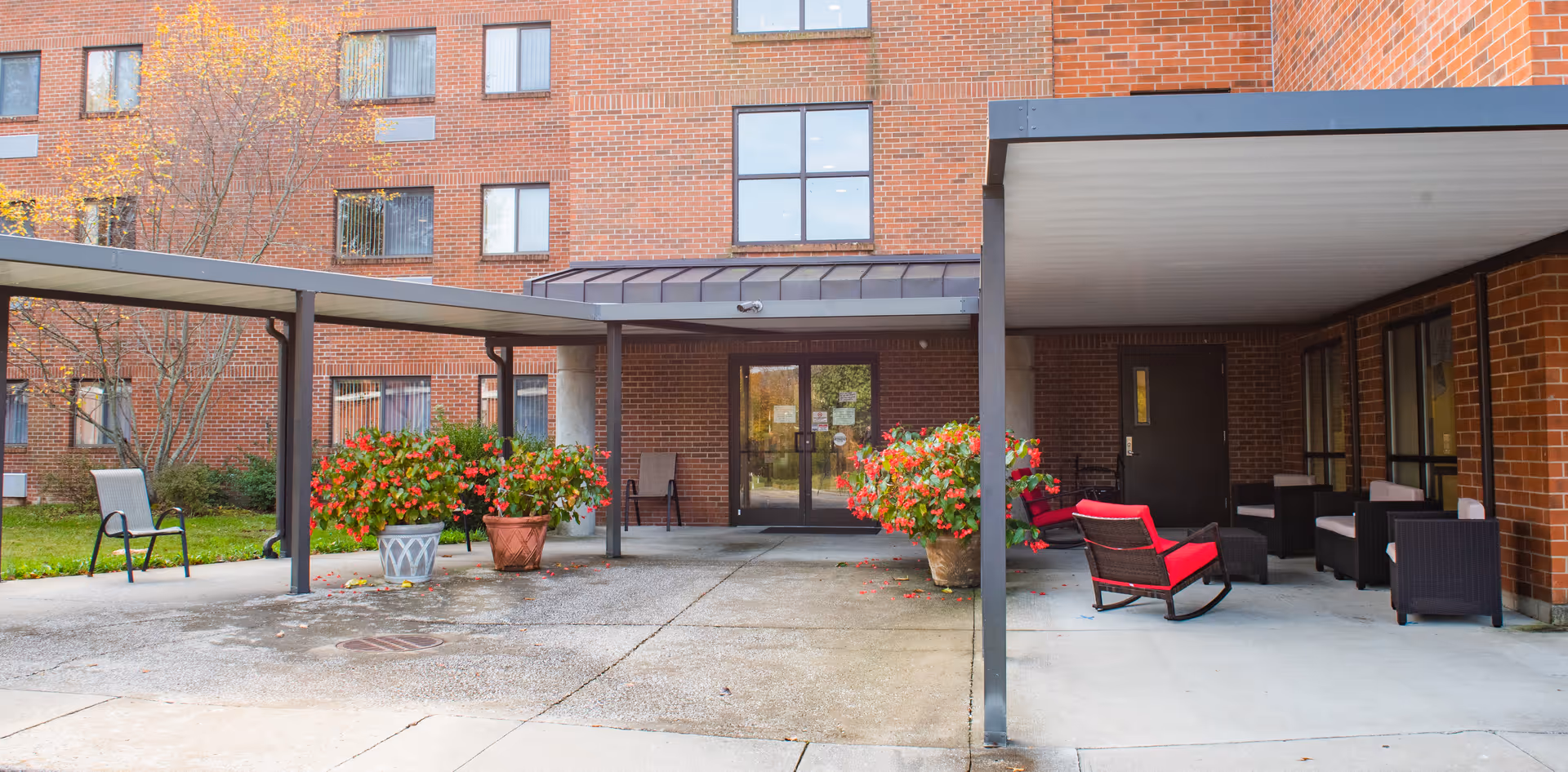 Entrance area of a brick senior living facility with a covered walkway and patio. There are potted plants with red flowers near the entrance, outdoor chairs including a red cushioned rocking chair, and a few other seating arrangements. The building has multiple windows and a glass door entrance.