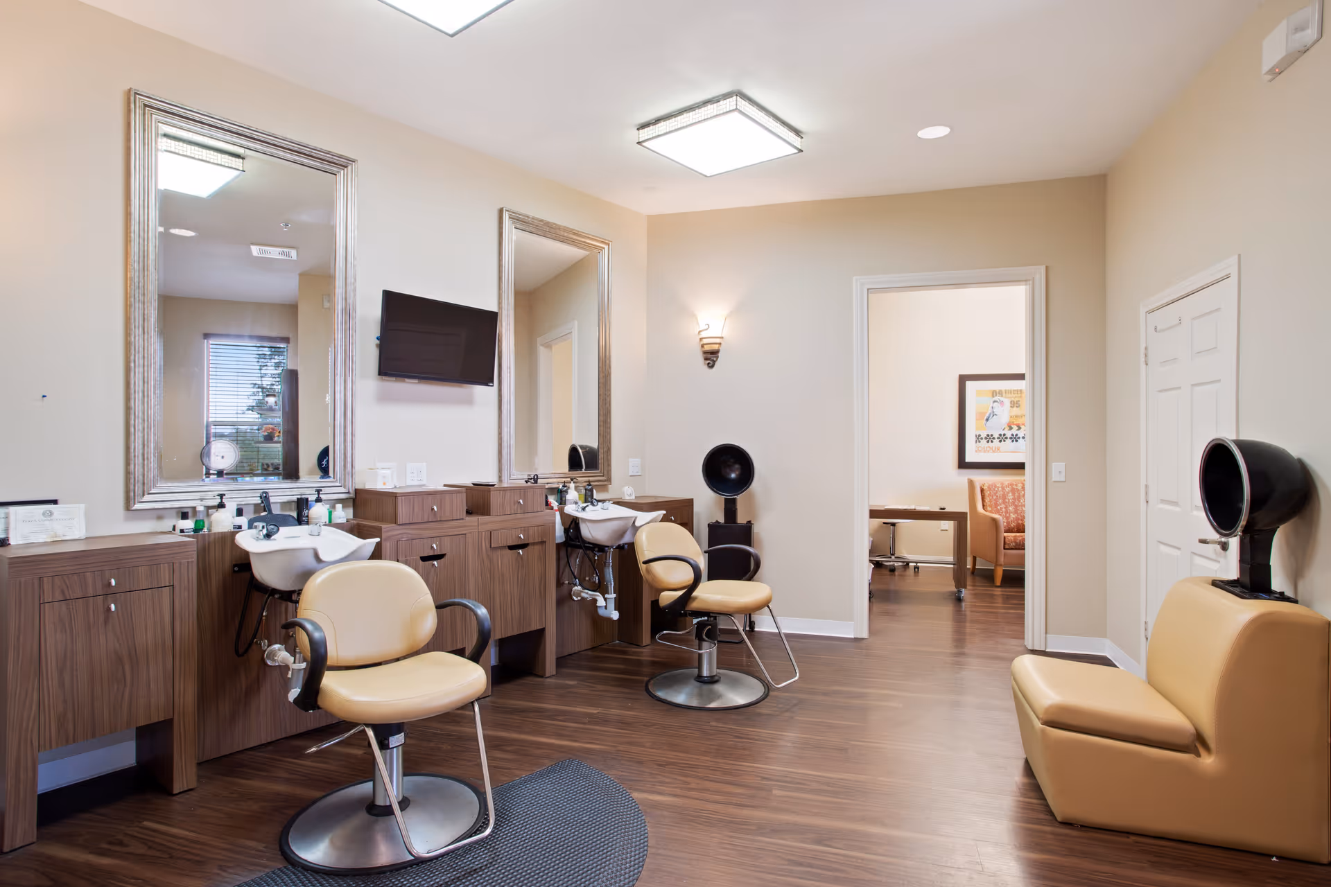 Interior view of a hair salon area in a senior living facility with two salon chairs in front of sinks and large mirrors, a wall-mounted TV, and a hair dryer chair. The room has wooden flooring and beige walls, with a doorway leading to another room with a desk and seating.