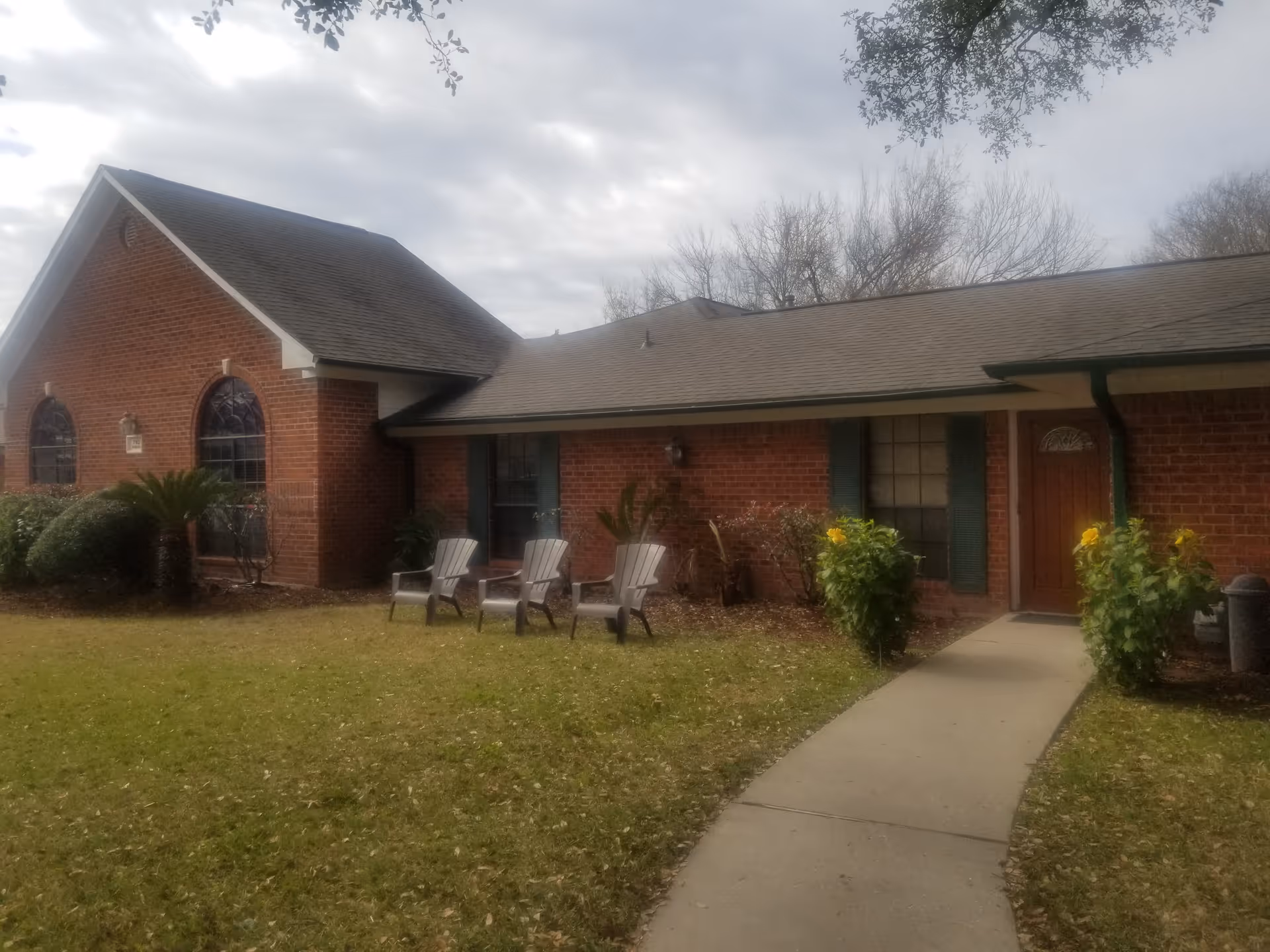 Exterior view of a single-story brick building with a sloped roof, featuring a walkway leading to a wooden door. There are three plastic chairs on the lawn near the building, surrounded by bushes and small plants. The sky is cloudy.