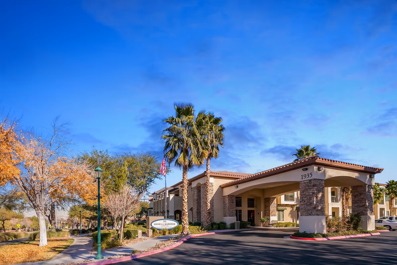 Front entrance of Merrill Gardens at Green Valley Ranch with a porte-cochère, palm trees, landscaped grounds, and a driveway.
