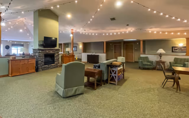 Open senior living common area with armchairs and tables arranged around a stone fireplace and wall-mounted TV under string lights.