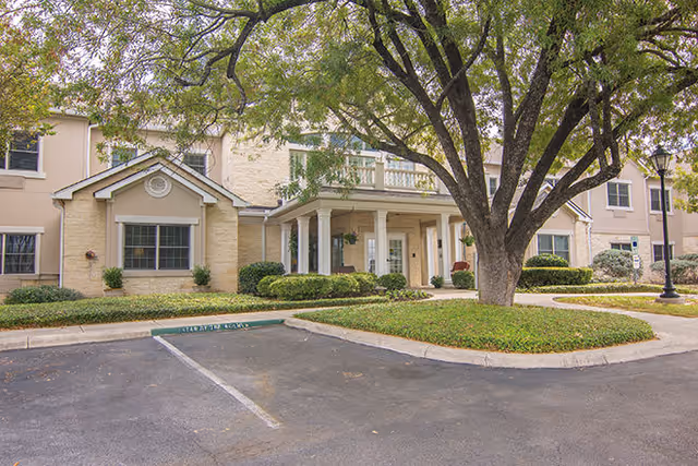 Exterior view of a senior living facility building with beige walls, multiple windows, and a covered entrance supported by white columns. There is a large tree with green leaves in front of the building, surrounded by landscaped bushes and grass. A parking area is visible in the foreground.