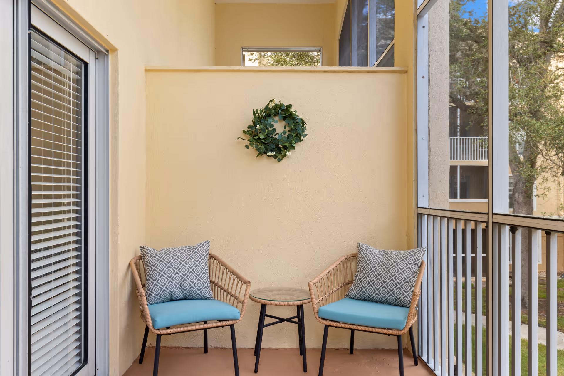 A small screened balcony with two wicker chairs featuring blue cushions and patterned pillows, a small round table between them, and a green wreath hanging on a beige wall. The balcony overlooks a courtyard with trees and other building balconies.