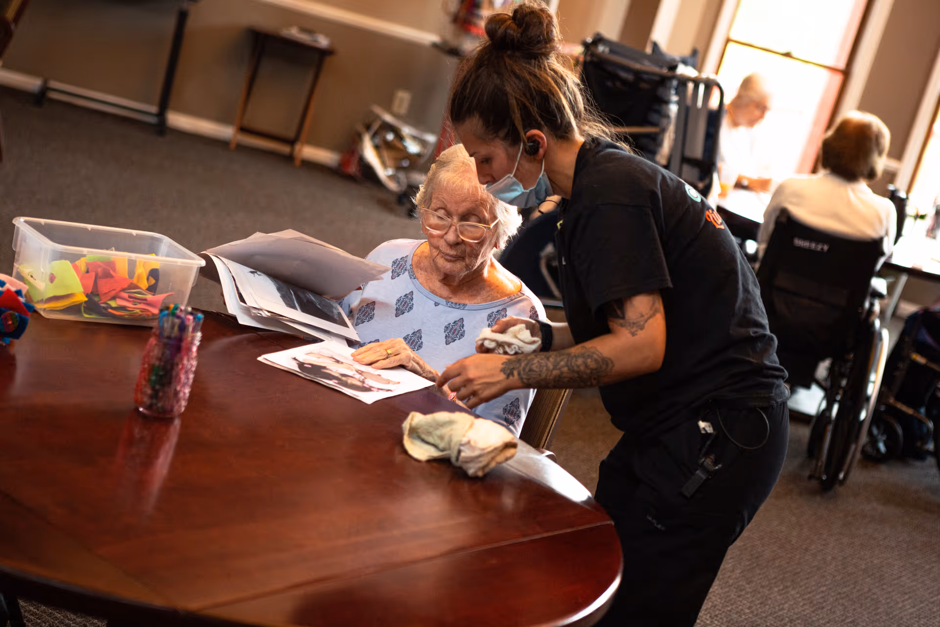 An elderly woman sitting at a wooden table looking at papers with a younger woman standing beside her, wearing a mask and assisting her. In the background, other elderly people are seated in wheelchairs near windows.