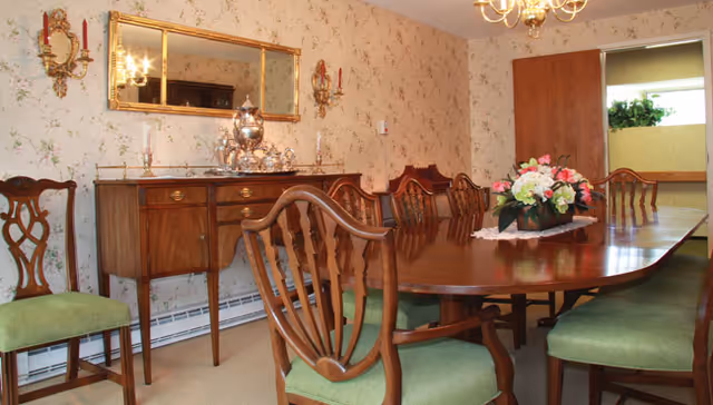 A traditional dining room with a long wooden dining table surrounded by wooden chairs with green cushions. A floral centerpiece is placed on the table. The room has floral wallpaper, a large mirror above a wooden sideboard with silver tea set, and two wall sconces with candles. A chandelier hangs from the ceiling.