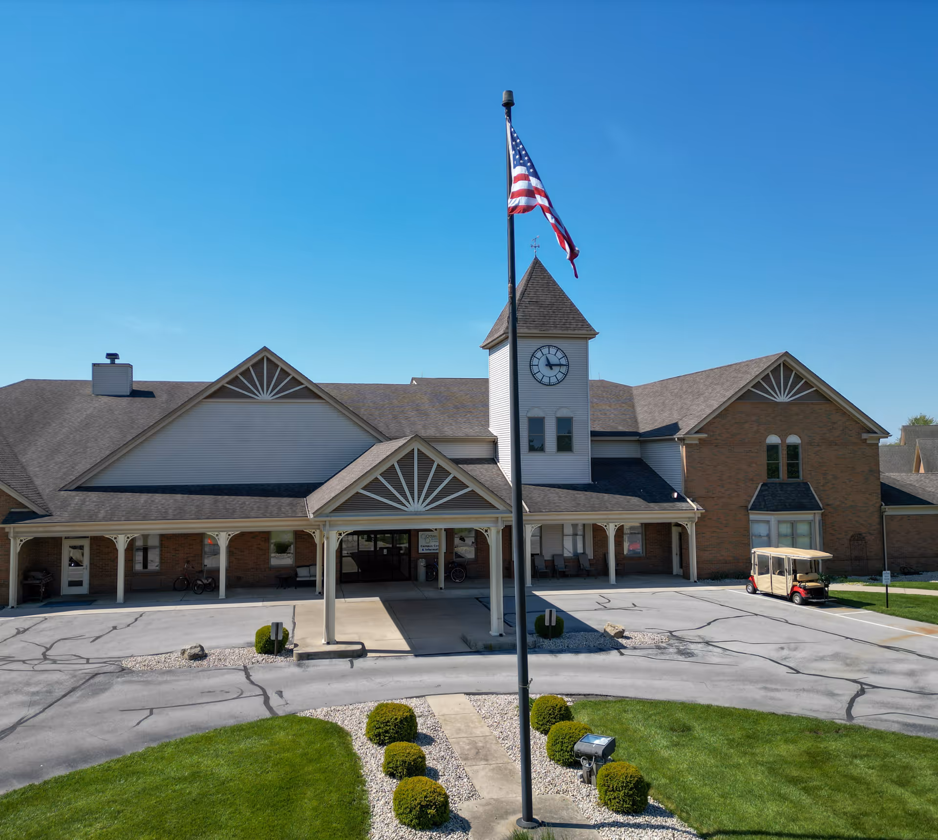 Exterior view of Otterbein St. Marys SeniorLife Community building with a clock tower and an American flag on a flagpole in front. The building has a covered entrance, brick and siding walls, and a golf cart parked on the right side. The sky is clear and blue.