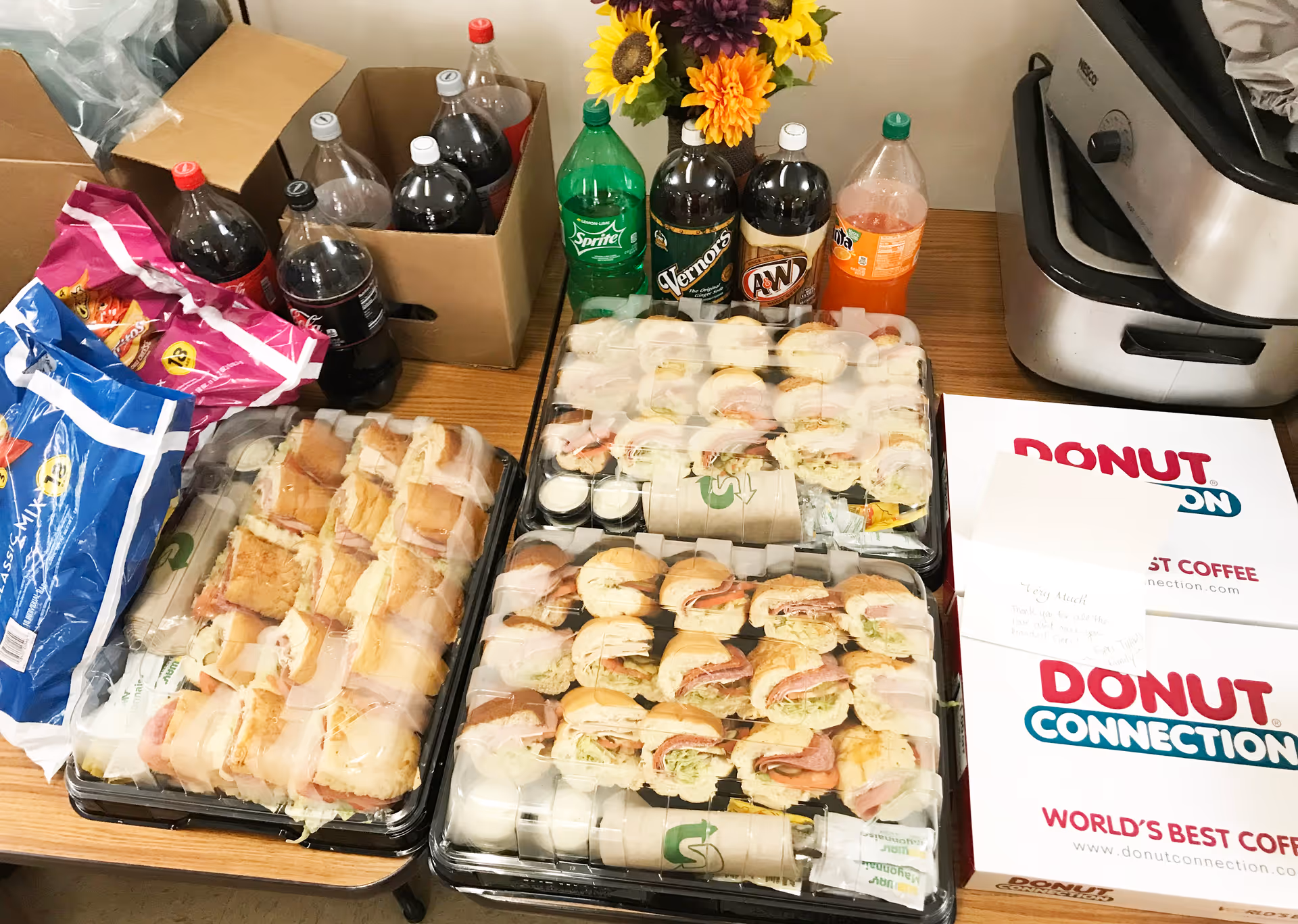 Table set with trays of assorted sandwiches, bottles of soda, snack bags and Donut Connection boxes.