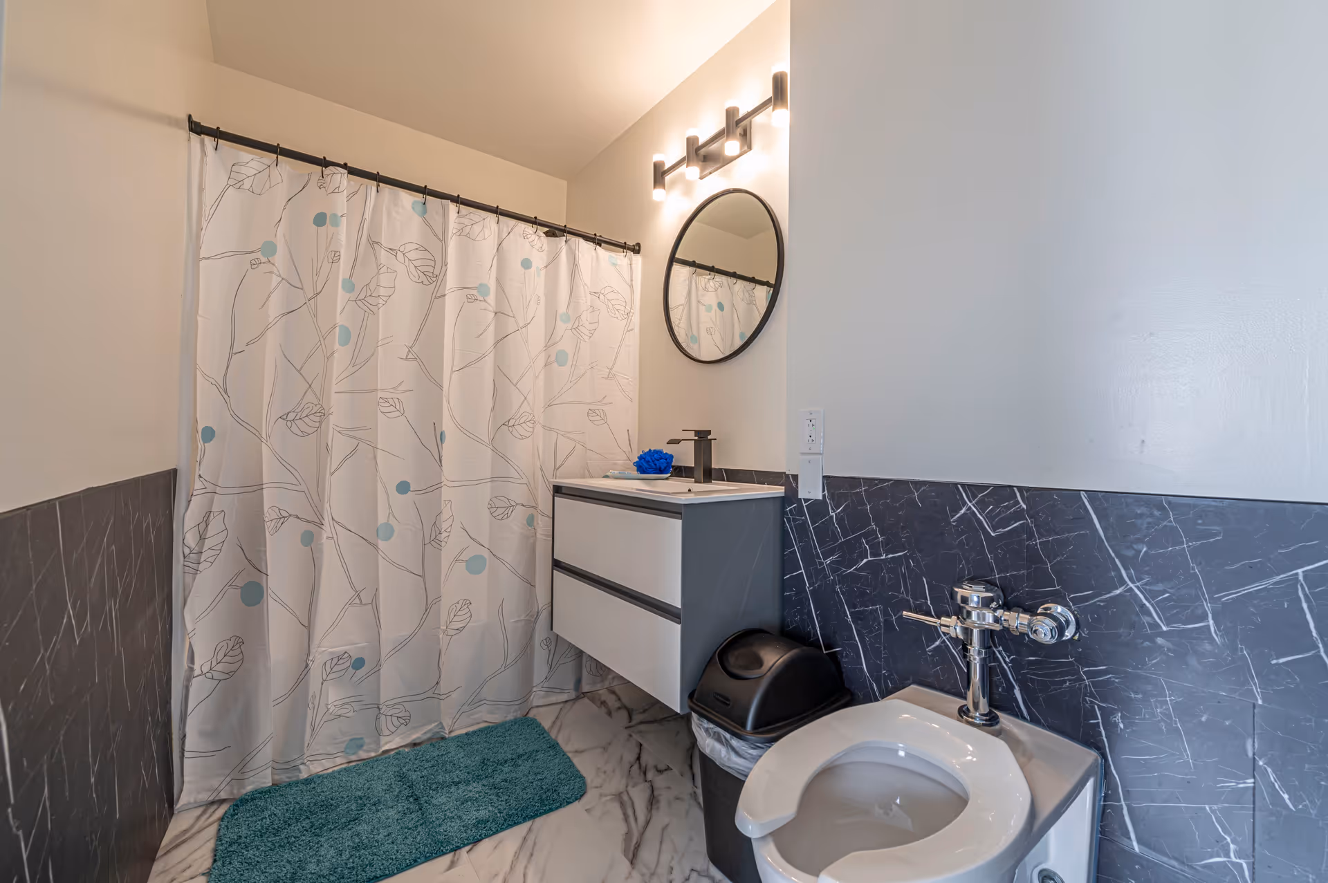 A modern bathroom with a white toilet, a black trash bin, a floating vanity with a sink, a round mirror above the sink, a light fixture with three bulbs, a shower curtain with a leaf and dot pattern, and a teal bath mat on a marble-patterned floor.