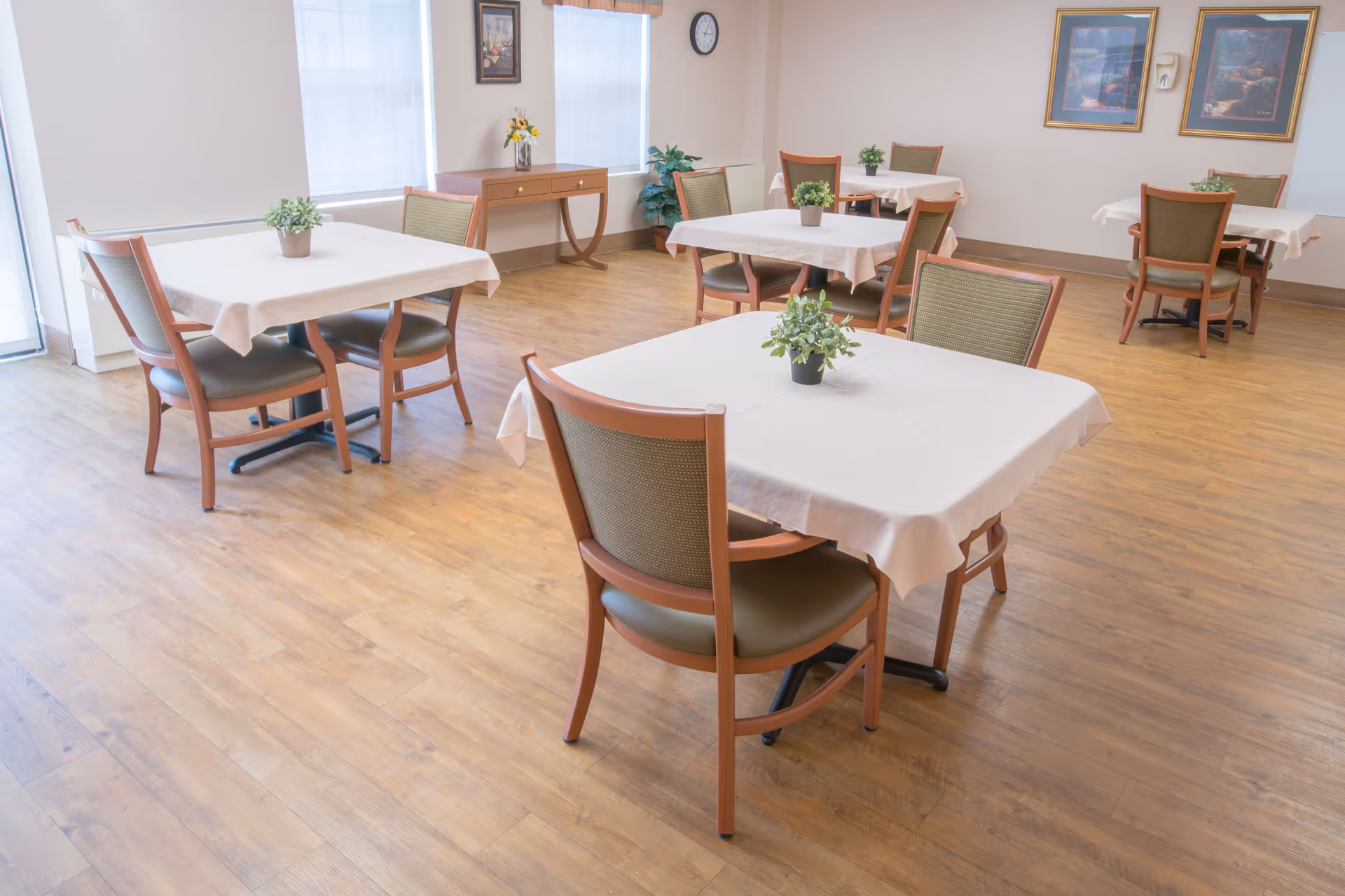 A bright dining room with wooden floors featuring several square tables covered with white tablecloths, each surrounded by four chairs with wooden frames and green cushions. Small potted plants are placed in the center of each table. The room has large windows with blinds, a wooden console table against the wall, and framed artwork hanging on the walls.