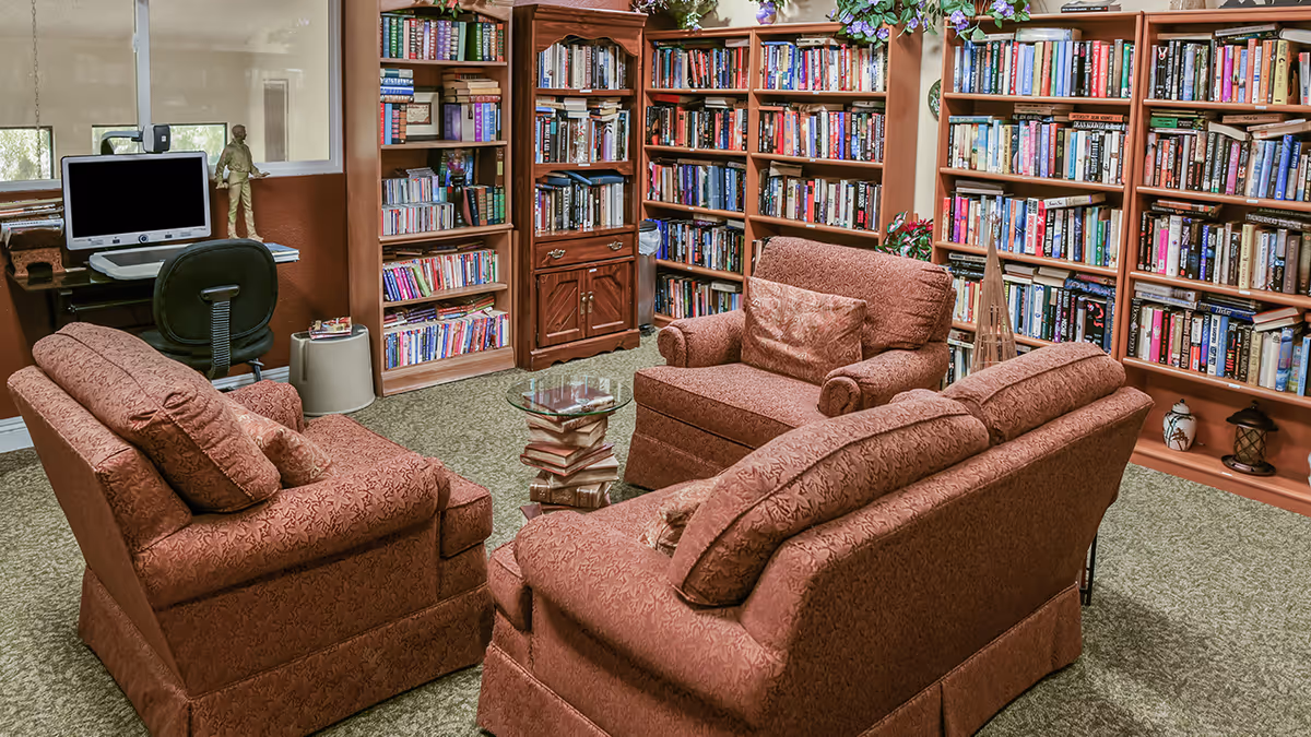A cozy library room with three upholstered armchairs and a loveseat arranged around a small glass-top table made of stacked books. The walls are lined with wooden bookshelves filled with books and decorative items. A computer workstation with a chair is visible in the corner near a window.