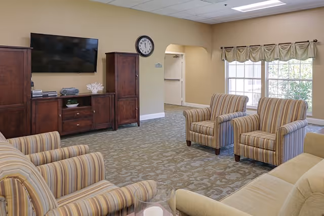 Well-lit common living room with striped armchairs and a sofa facing a wall-mounted TV and wooden media console near large windows.