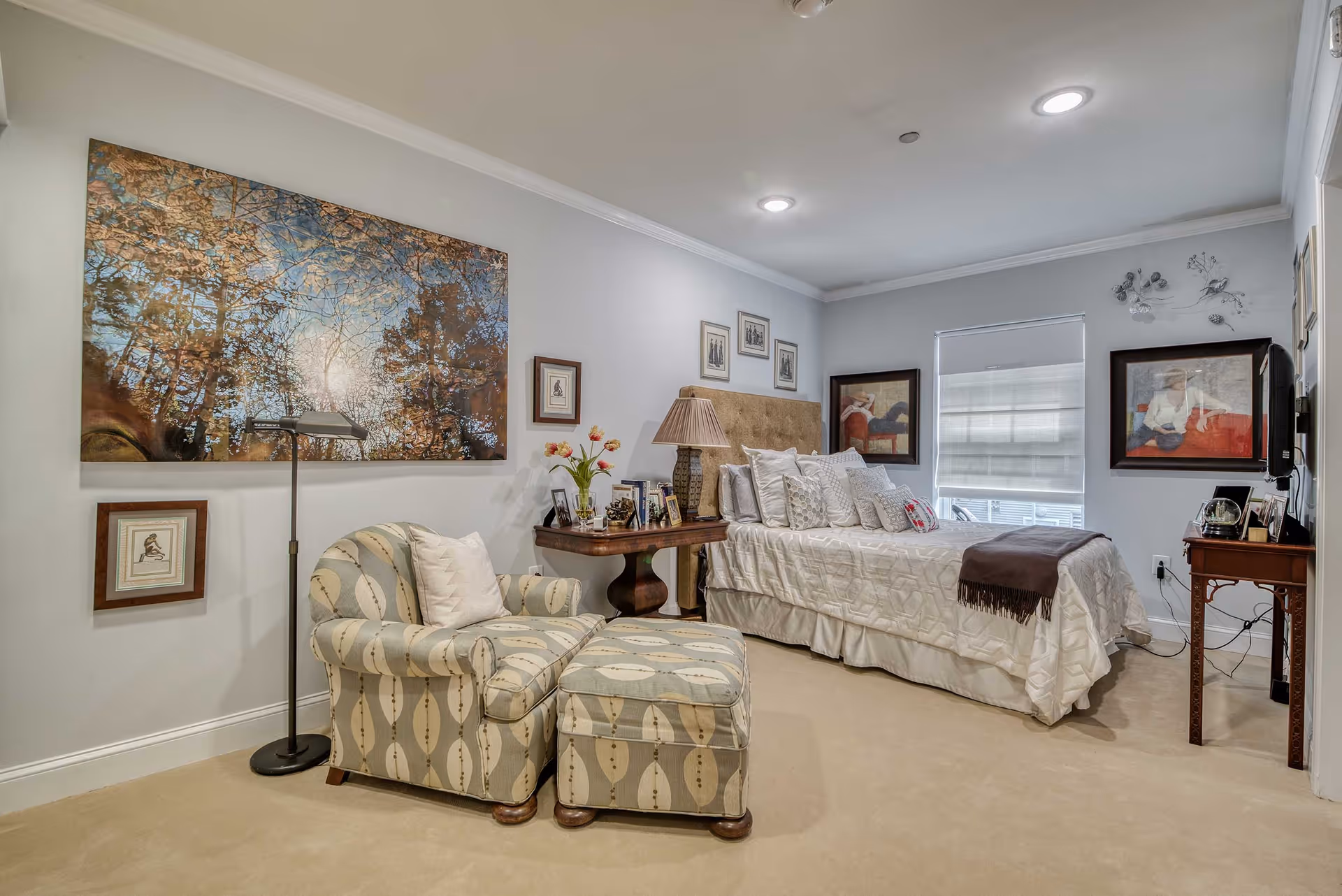 Well-lit, decorated bedroom with a bed, patterned armchair and ottoman, side tables, and framed artwork on the walls.