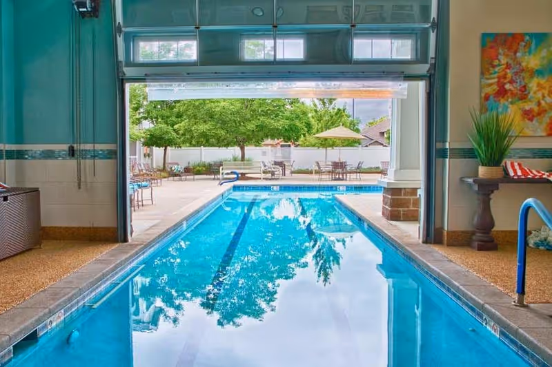 Indoor lap pool with a roll-up door open to an outdoor patio area with tables, umbrellas and trees.