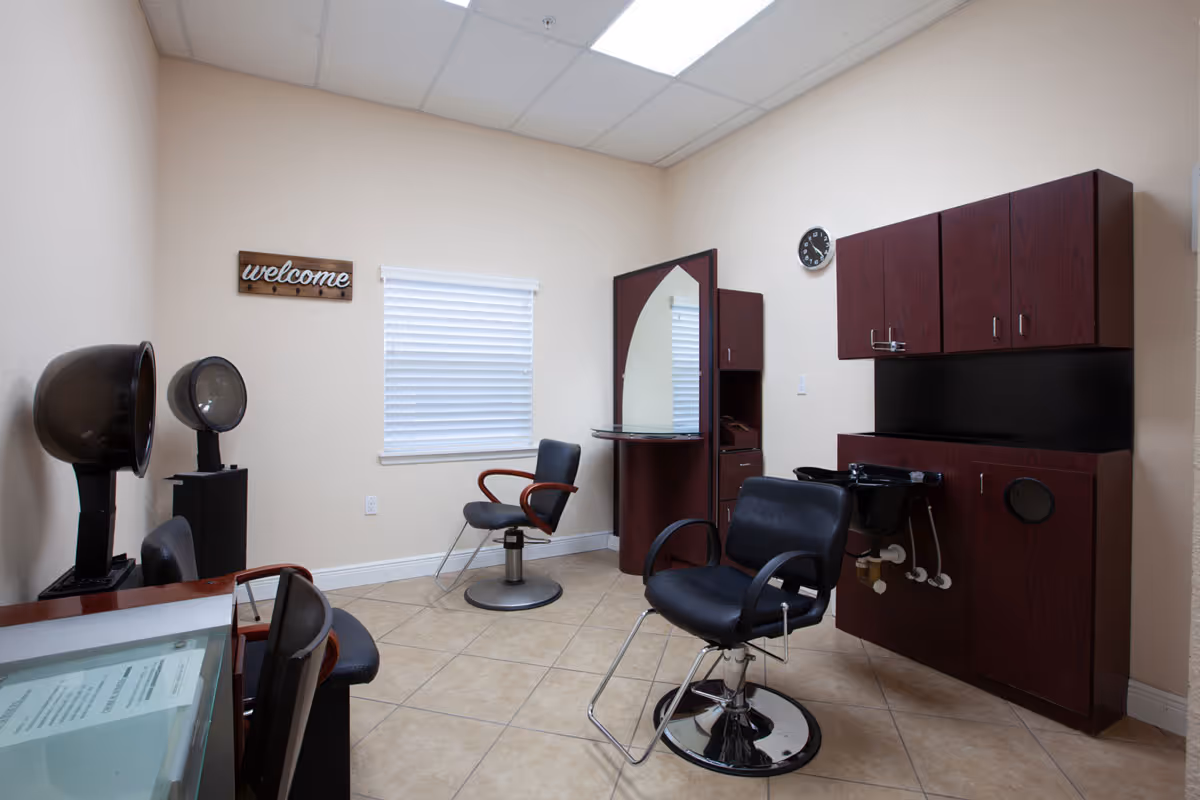 Small salon-style room with barber chairs, hooded hair dryers, a mirror station, and wall cabinets beneath a 'welcome' sign.