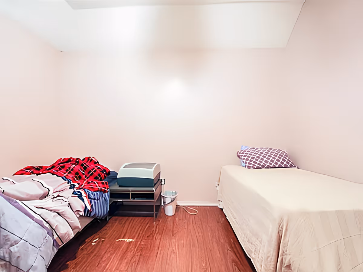 A small bedroom with two single beds on opposite sides of the room. One bed has a red and black checkered blanket and other bedding, while the other bed has a plain beige cover and a purple patterned pillow. There is a small nightstand between the beds with a device on it and a small trash can on the floor. The walls are light-colored and the floor is wooden.