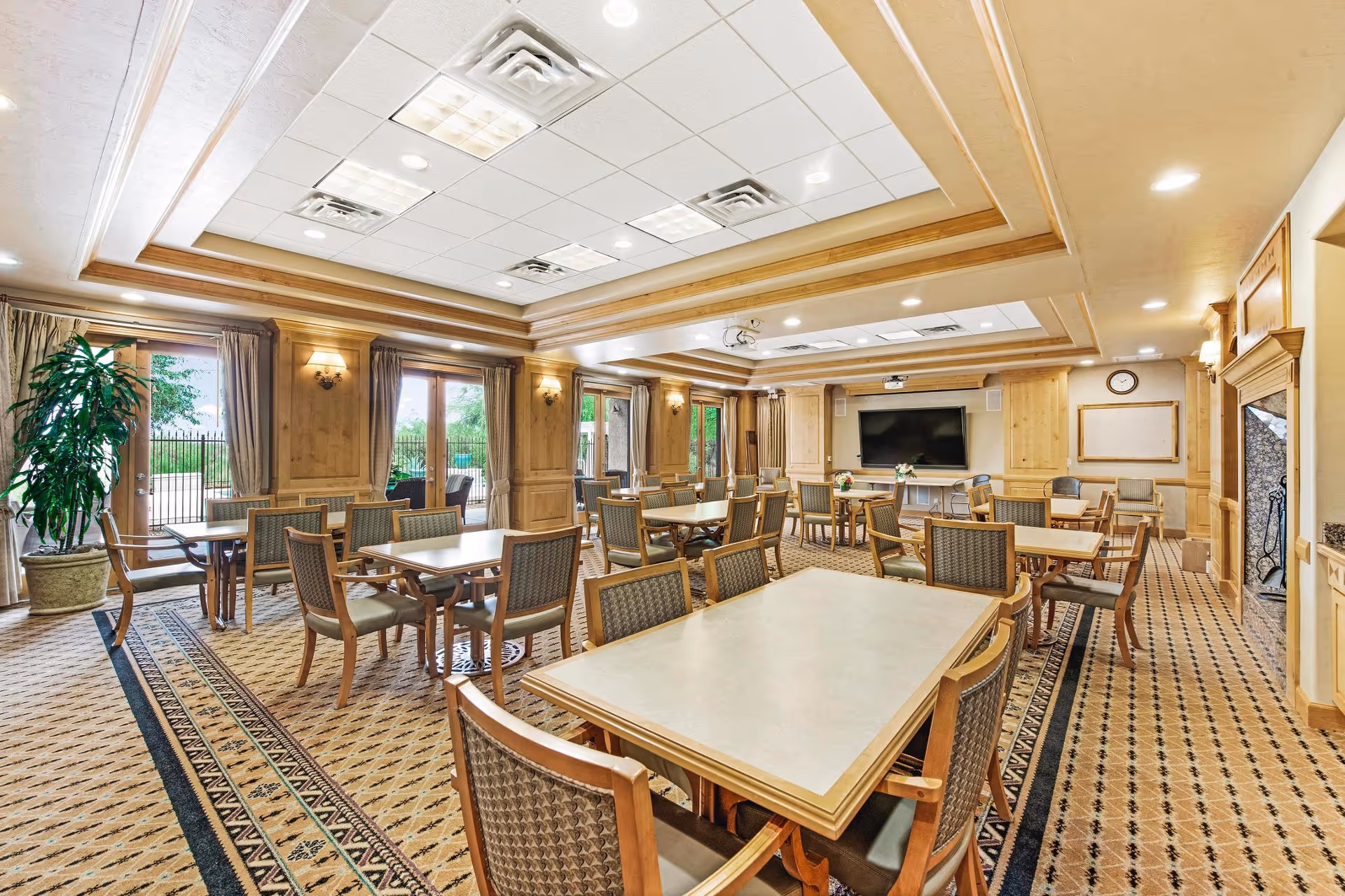 Well-lit communal dining/activity room with multiple tables and chairs, a large TV, and windows looking outside.