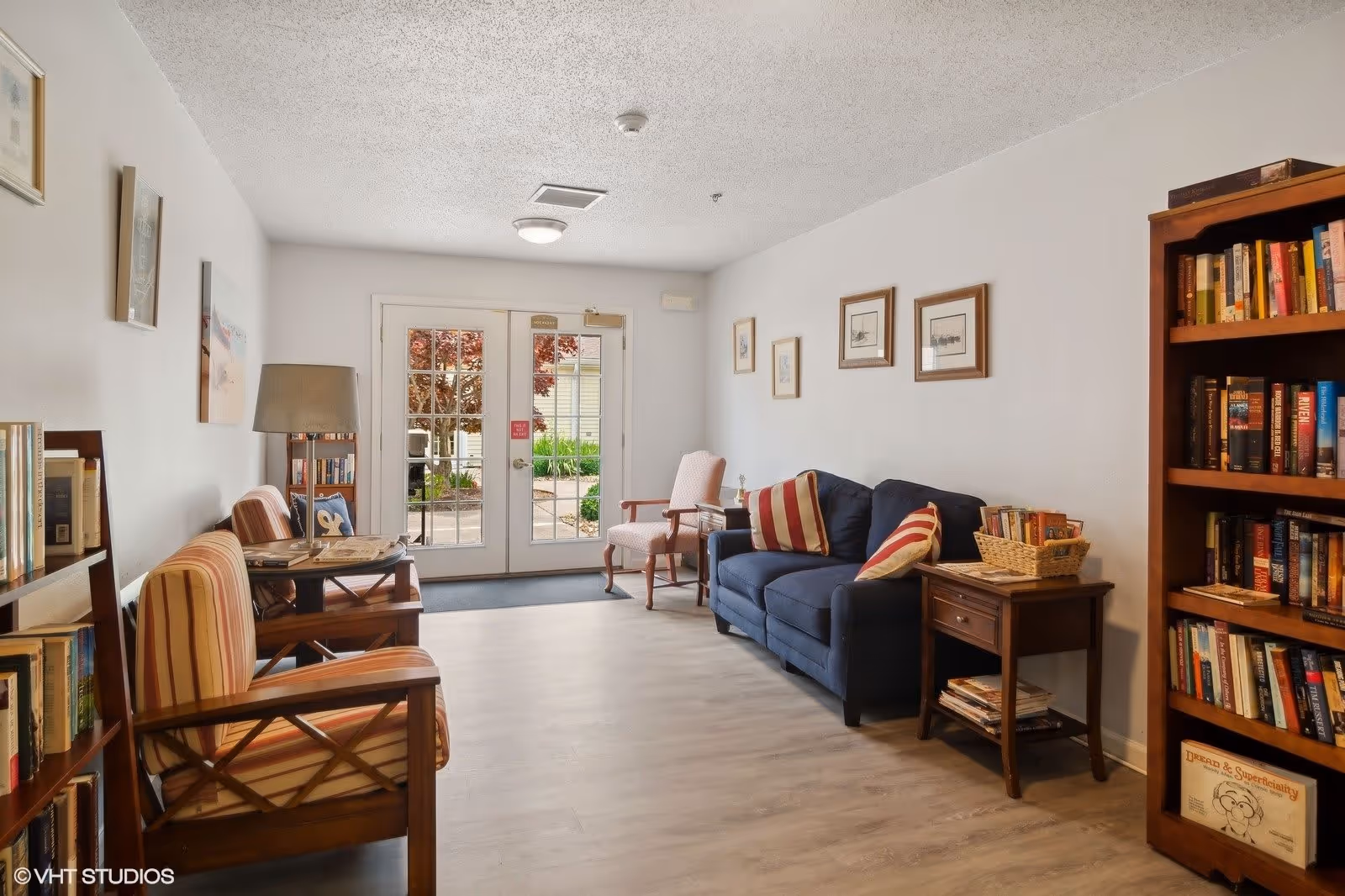 A cozy sitting area in a senior living facility with a blue couch adorned with striped pillows, two striped armchairs, a wooden side table with magazines, and bookshelves filled with books. The room has light-colored walls decorated with framed pictures and a glass door leading outside, allowing natural light to brighten the space.