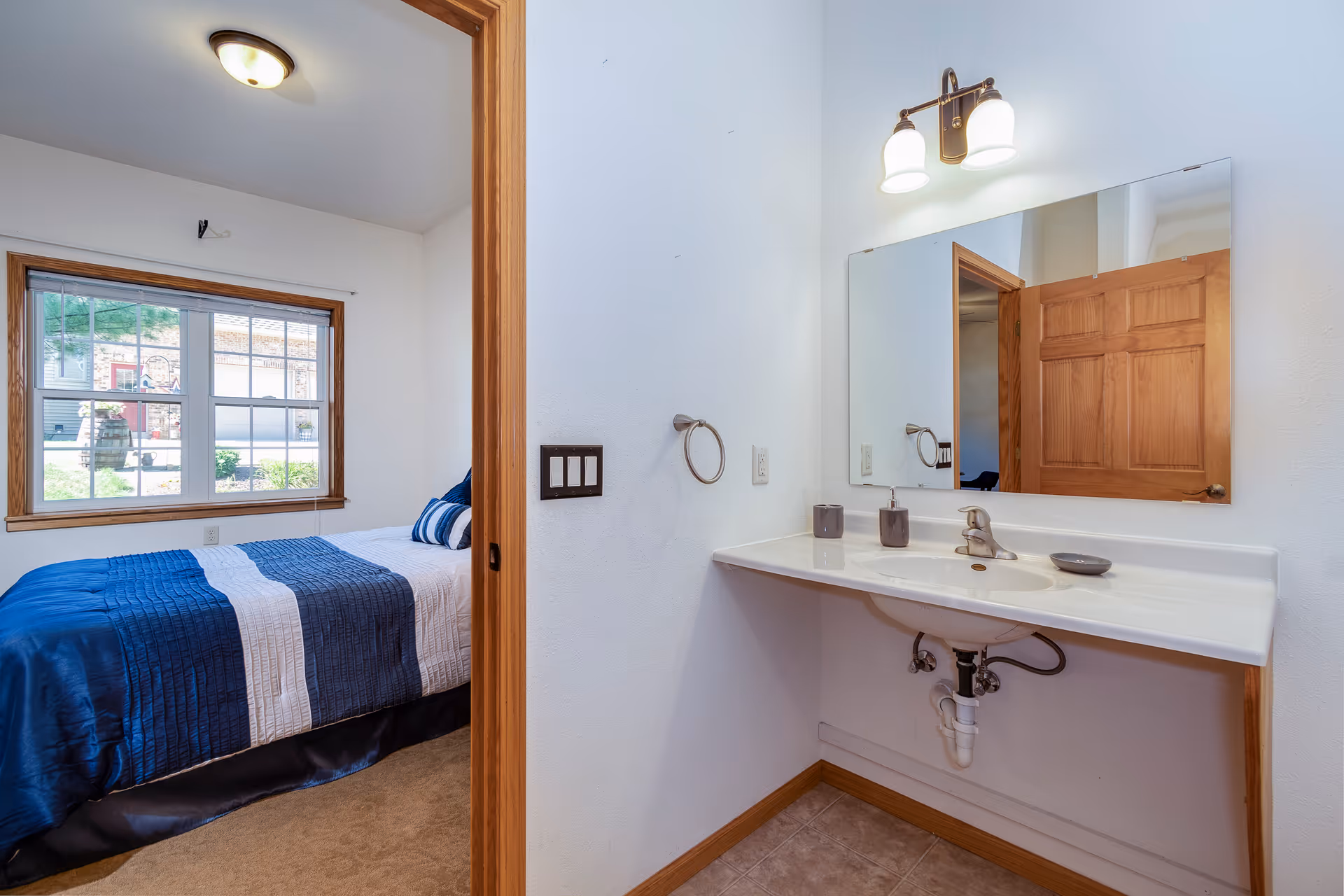 View of a bedroom with a blue-striped bed seen through a doorway beside a bathroom sink, mirror, and light fixture.