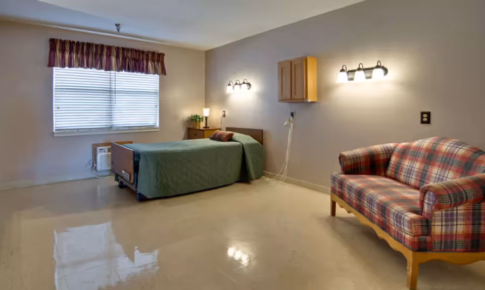 A simple senior living bedroom with a single bed, nightstand, wall-mounted lights, and a plaid loveseat.