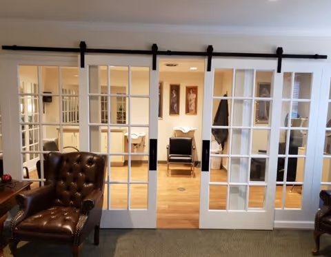 Interior view of a senior living facility room with white sliding glass doors. In front of the doors are two brown leather tufted armchairs. Through the doors, there are two black chairs with white wash basins behind them, and framed artwork on the walls. The floor is wooden and the room is well-lit.