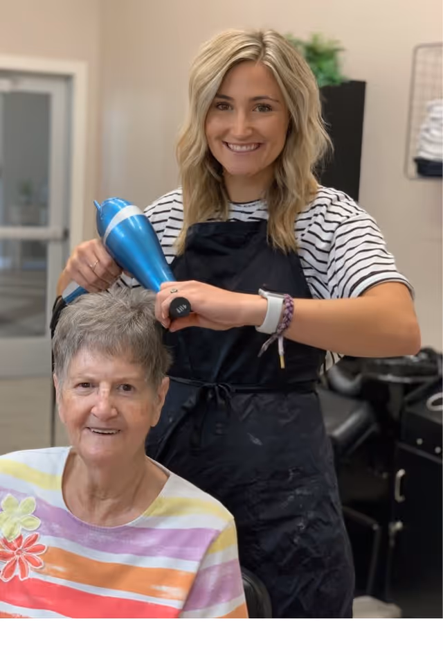 A smiling hairstylist with blonde hair is blow-drying the short gray hair of an elderly woman who is also smiling. They are in a salon setting with hairdressing equipment visible in the background.