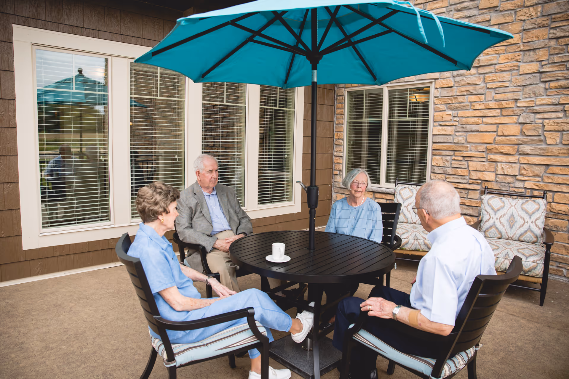 Four elderly people sitting around a round outdoor table with a large blue umbrella, on a patio area next to a building with stone and wood siding. There are cushioned chairs and a bench with patterned cushions in the background.