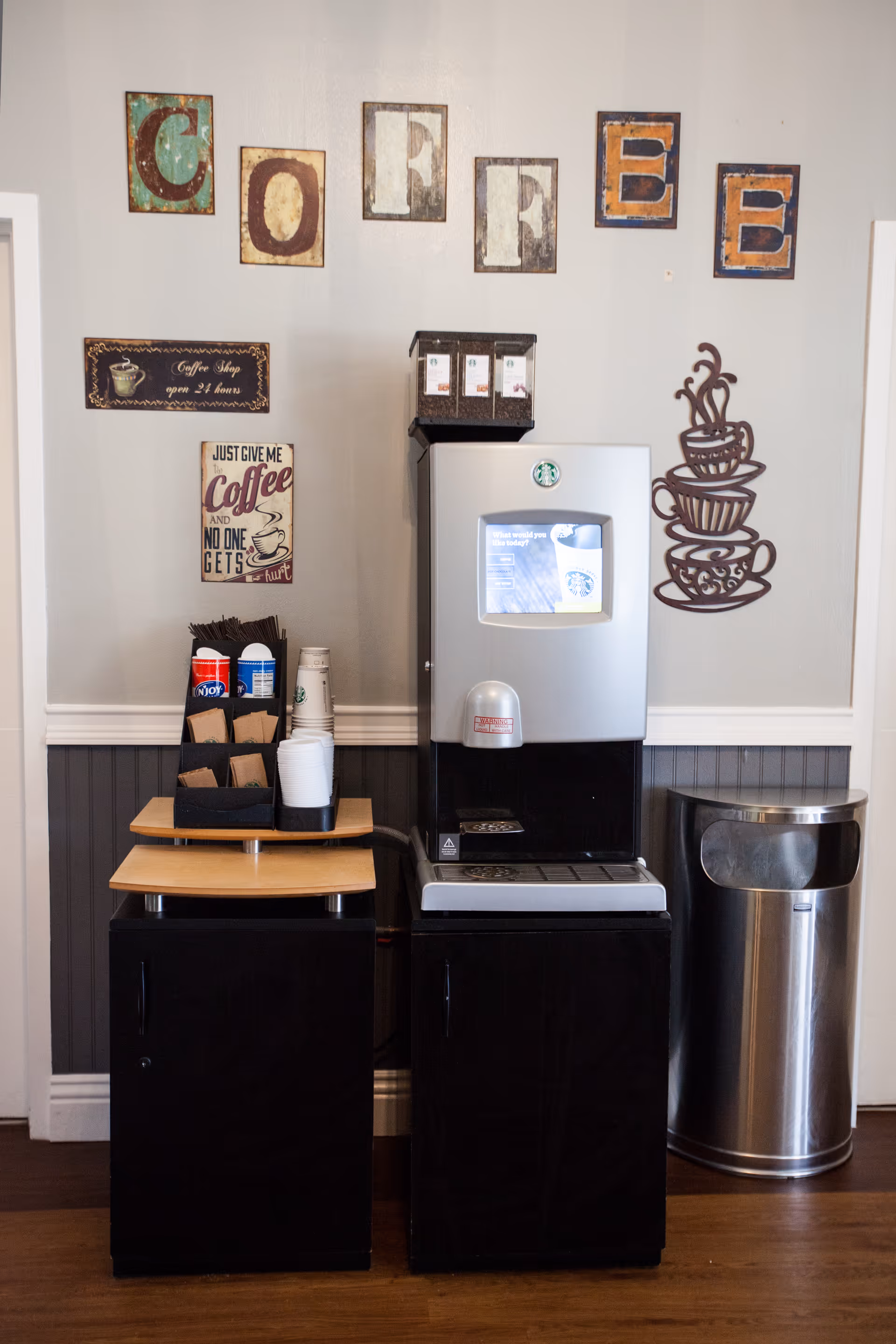 A coffee station with a modern coffee machine, disposable cups, stirrers, and coffee condiments on a small wooden counter. The wall behind features decorative signs spelling out 'COFFEE' and coffee-themed artwork. A stainless steel trash can is placed to the right of the station.