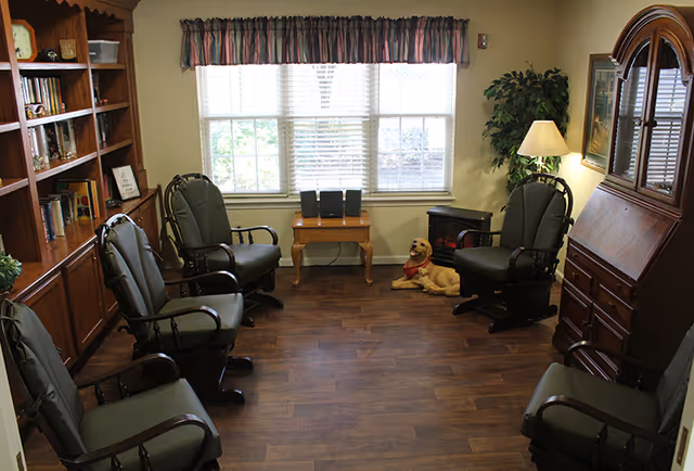 A cozy living room with six cushioned chairs arranged in a semi-circle on a wooden floor. There is a wooden bookshelf filled with books and decorative items on the left, a small wooden table with three black speakers in front of a window with blinds and a valance. A lamp and a potted plant are in the corner, and a decorative dog statue is lying on the floor near a small electric fireplace. A wooden cabinet with glass doors is on the right side of the room.