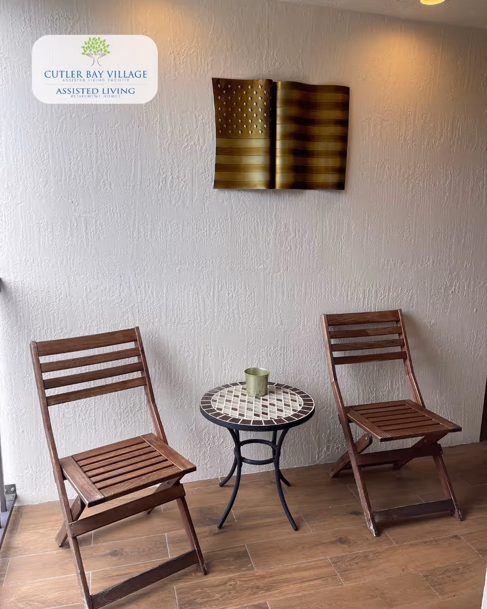 Two wooden folding chairs with slatted seats and backs are positioned on either side of a small round mosaic table with a metal base. A small metallic cup is placed on the table. The setting is indoors against a textured white wall with a decorative metal American flag mounted on it. The floor is covered with wood-like tiles.