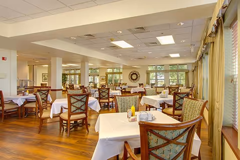 A spacious dining room with multiple tables covered with white tablecloths and wooden chairs with patterned cushions. The room has large windows with curtains allowing natural light to fill the space, and the floor is made of polished wood. The ceiling has recessed lighting and air vents, and there are decorative elements on the walls.
