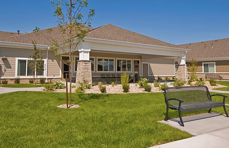 Outdoor view of Sunridge Assisted Living of Roy showing a well-maintained lawn with small trees and shrubs, a paved walkway, a black metal bench, and a beige building with a covered porch and multiple windows under a clear blue sky.