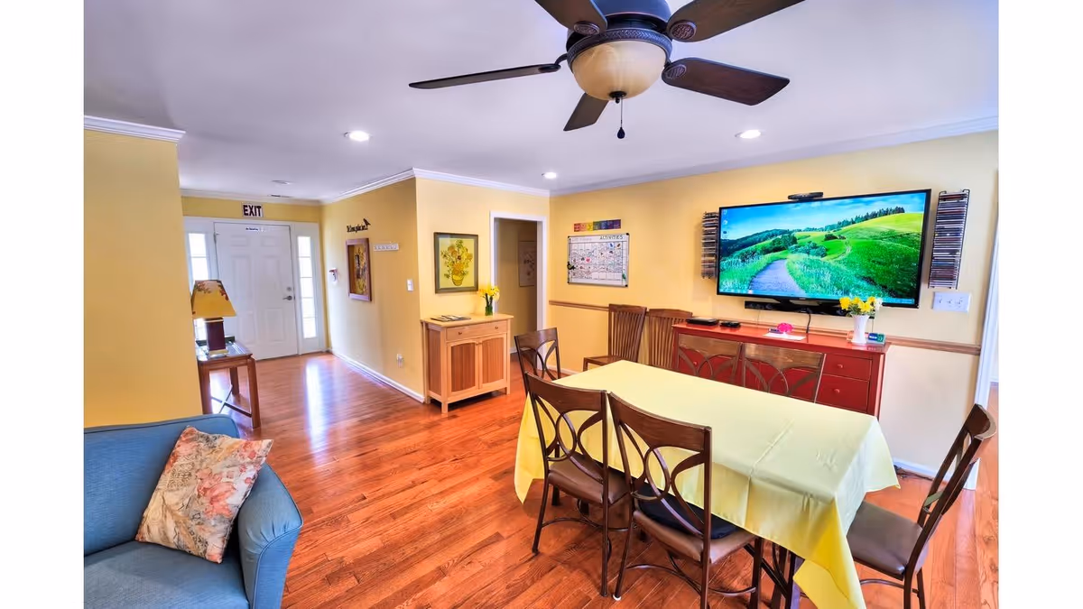 A bright and cozy common area with yellow walls and wooden flooring. There is a rectangular table covered with a yellow tablecloth surrounded by six wooden chairs. A large flat-screen TV is mounted on the wall above a red cabinet, displaying a scenic landscape. A ceiling fan with lights is centered on the white ceiling. To the left, there is a blue armchair with a floral cushion, a small table with a lamp, and a white door marked with an exit sign. The room is decorated with framed pictures and a calendar on the walls.