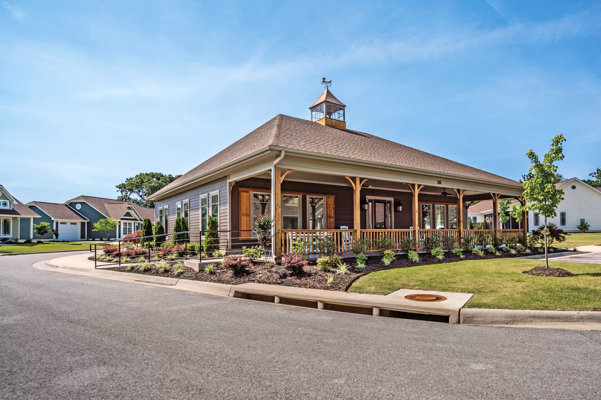 Exterior view of a single-story building with a wraparound porch, wooden railings, and a cupola on the roof. The building is surrounded by landscaped greenery and a paved road curves around it. Other similar residential buildings are visible in the background under a clear blue sky.