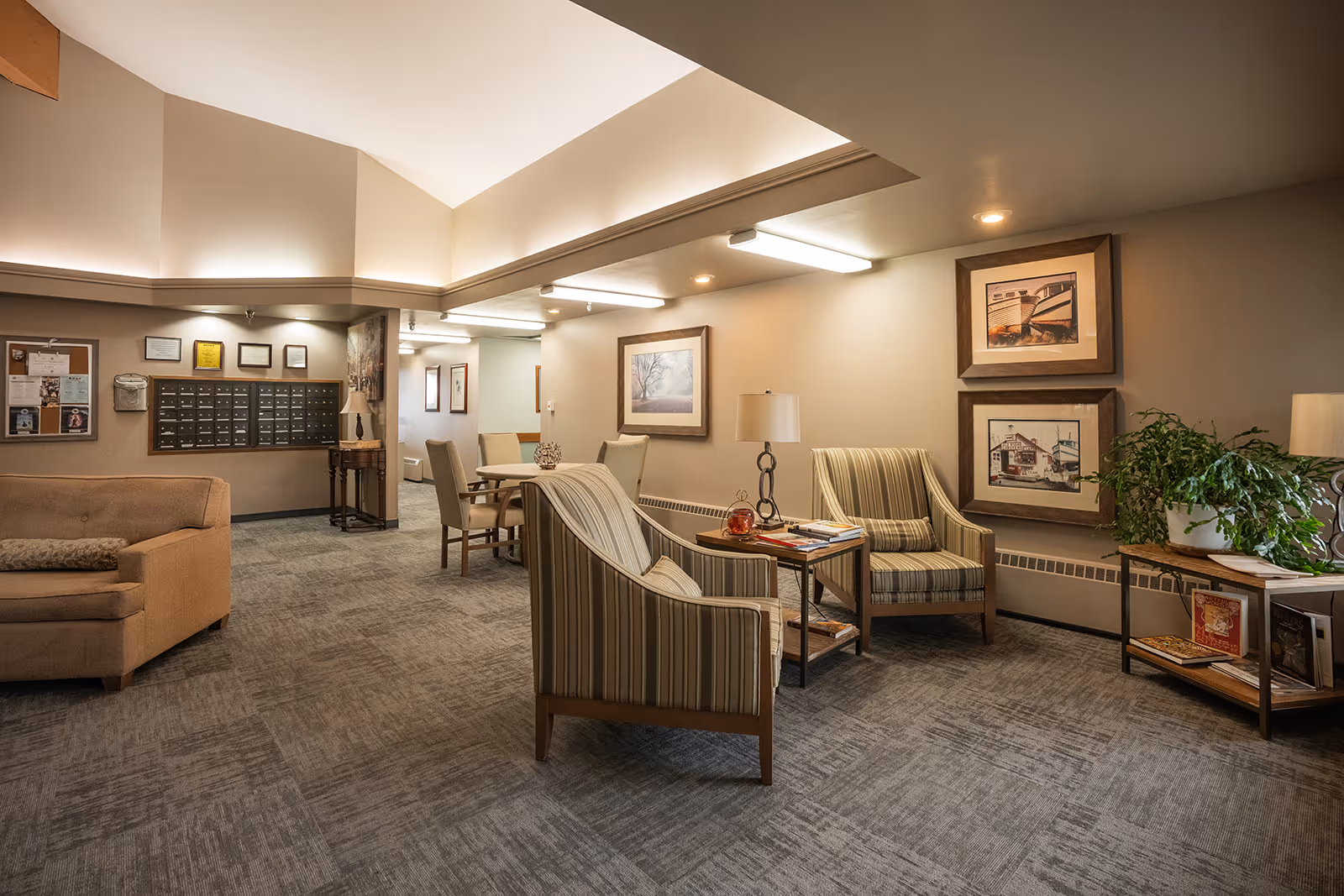 Well-lit communal lounge with armchairs, a small table, mailboxes and framed artwork on the walls.