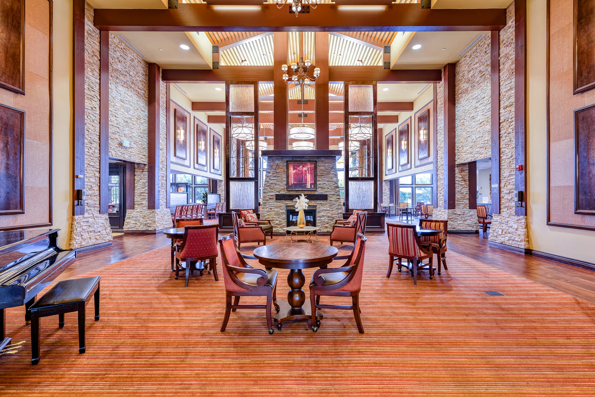 Spacious senior living common room with high ceilings, a central stone fireplace, round tables and chairs, and a piano.