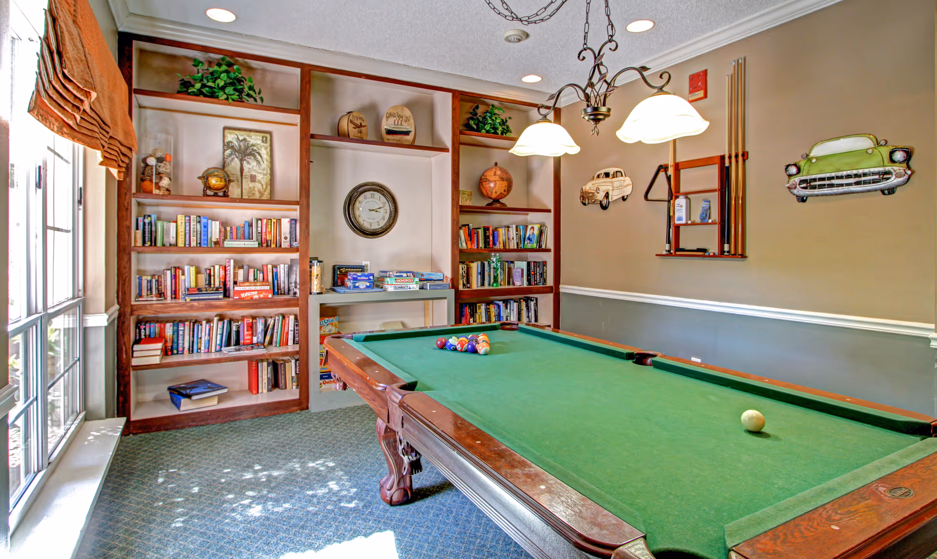 A recreational room with a green felt pool table in the center, surrounded by built-in wooden bookshelves filled with books and board games. The room has beige walls with decorative car-themed wall hangings and a rack holding pool cues. Two hanging pendant lights illuminate the pool table. A large window with a brown roman shade allows natural light into the room.