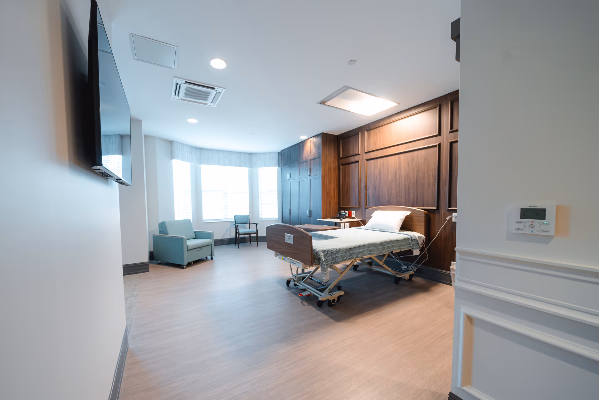 A spacious and well-lit bedroom in a senior living facility with a hospital-style bed, a wooden paneled wall behind the bed, a flat-screen TV mounted on the wall, a green armchair, a wooden chair, and large windows with blinds allowing natural light to fill the room.