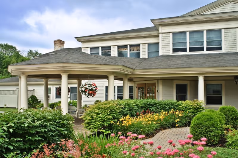Front entrance of a senior living building with a covered porte-cochère, columns, and landscaped flower gardens.