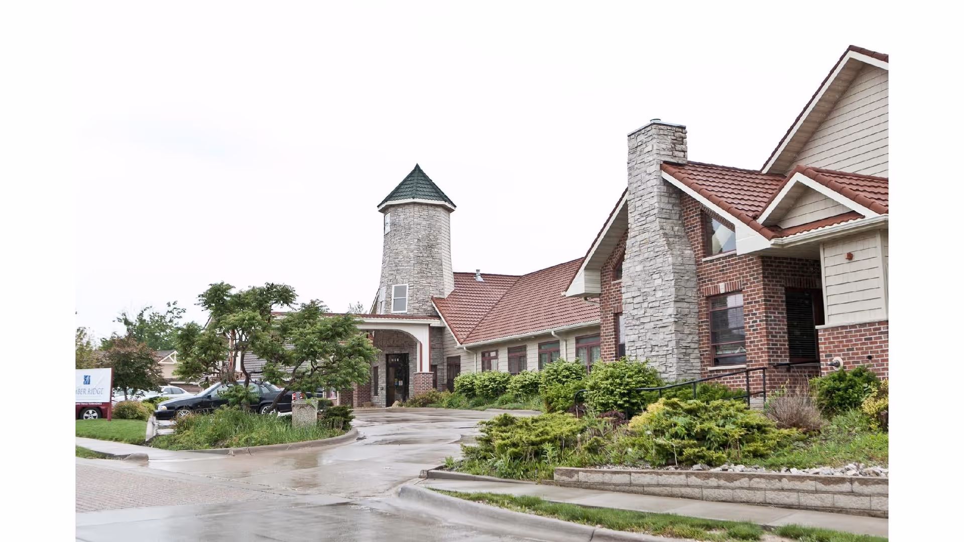 Exterior view of a senior living facility building with a brick and stone facade, a tower-like structure with a green roof, and a red-tiled roof. There is a driveway leading to the entrance, surrounded by greenery and small trees.