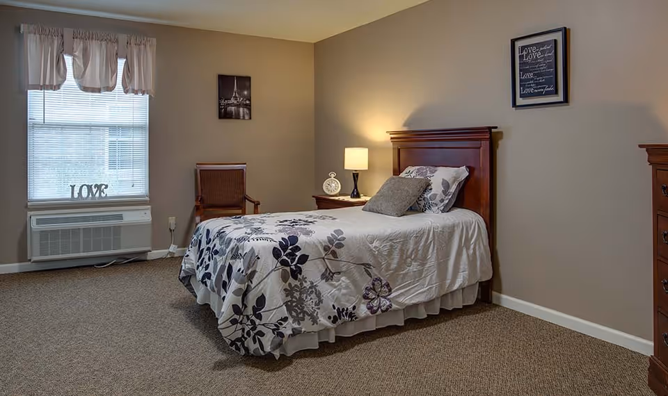 A tidy bedroom with a single bed in floral bedding, a nightstand and lamp, a wooden chair, and a window with 'LOVE' decor.