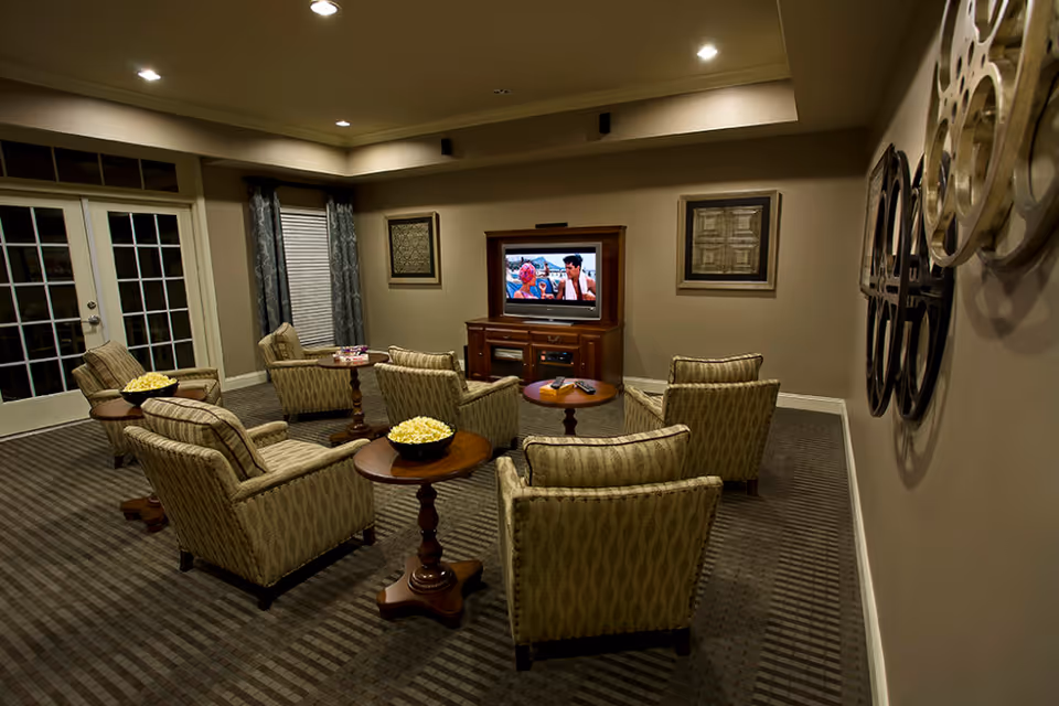 A cozy living room with six patterned armchairs arranged in two rows facing a wooden TV stand with a television showing a scene from a movie. There are small round wooden tables between the chairs, some with bowls of popcorn. The room has beige walls, framed artwork, a window with curtains, and French doors on the left side.
