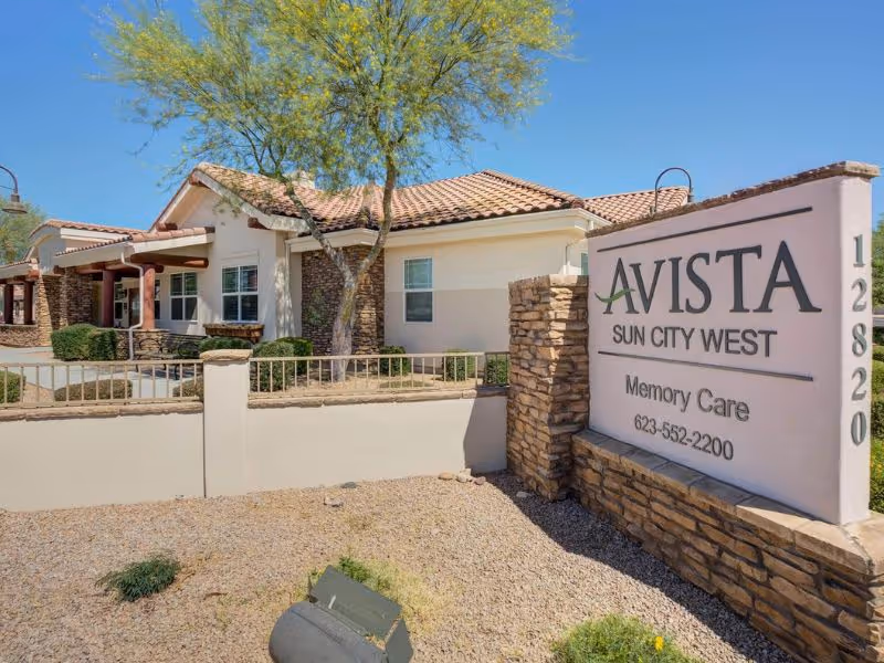 Exterior view of Avista Sun City West Memory Care facility showing a single-story building with a tiled roof, stone and stucco walls, a tree in front, and a large sign displaying the facility name, memory care designation, phone number, and address number 12820.
