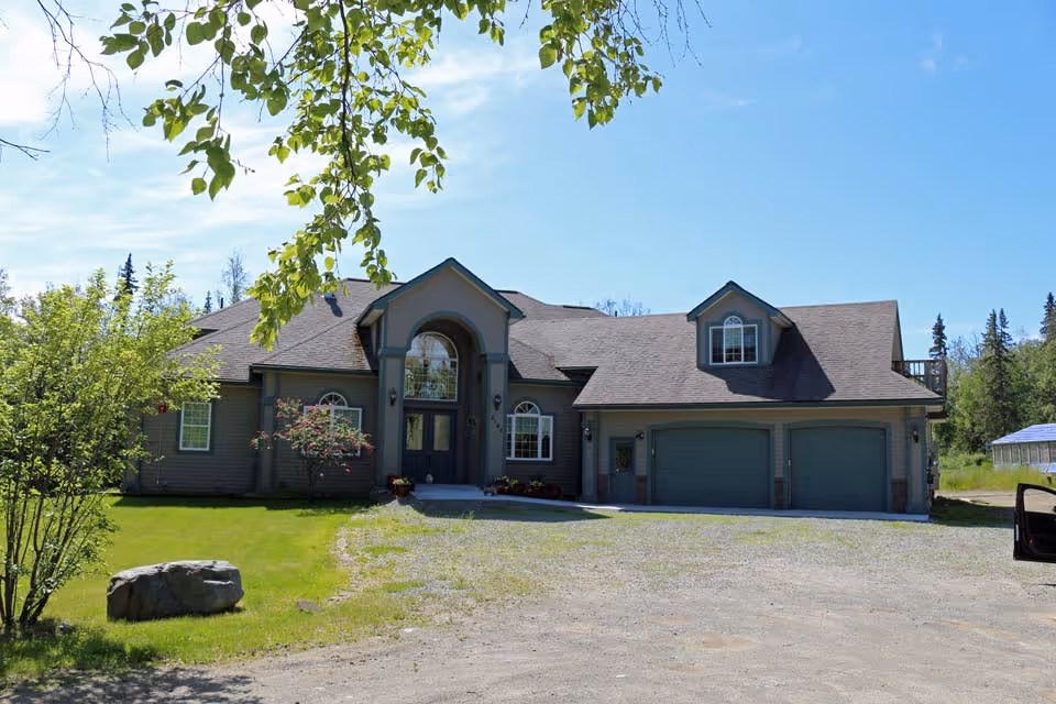 A large two-story house with a gray exterior, multiple windows, and a double garage under a clear blue sky. The house is surrounded by green grass, trees, and a gravel driveway.