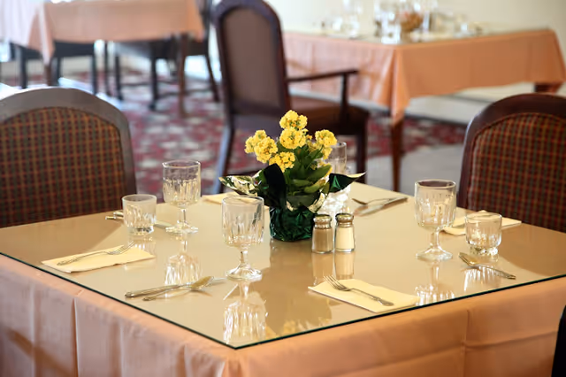 A dining table set for four with glassware, silverware, napkins, salt and pepper shakers, and a small potted yellow flower centerpiece. The table has a glass top and a peach-colored tablecloth. In the background, other similarly set tables and chairs are visible.